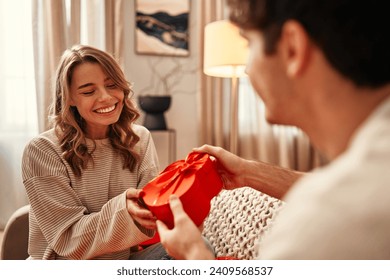 Happy Valentine's Day. Young couple in love exchanging gifts while sitting on the sofa in the living room at home. A man giving a heart-shaped gift box to his beloved woman. - Powered by Shutterstock