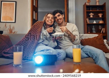 Happy Valentine's Day. A young couple in love sitting on the sofa in the living room at home, eating popcorn, covered with a blanket and watching a movie on the projector.