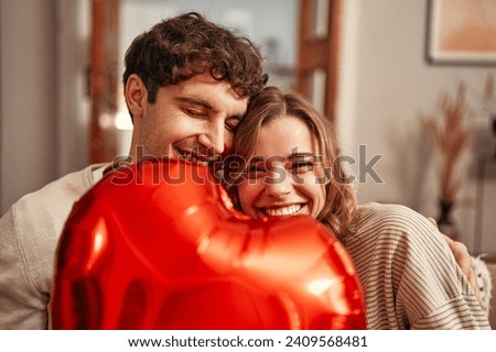 Photo of Happy Valentine's Day. Young couple in love holding a heart-shaped balloon, hiding behind it while sitting on the sofa in the living room at home. Romantic evening together.