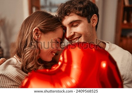 Happy Valentine's Day. Young couple in love holding a heart-shaped balloon while sitting on the sofa in the living room at home. Romantic evening together.