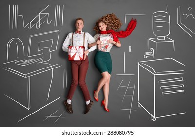 Happy Valentines Love Story Concept Of An Office Romance. Young Couple At Work Smiling At Each Other And Sharing Presents Against Chalk Drawings Background.