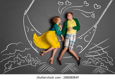Happy valentines love story concept of a romantic couple swinging on the moon, holding hands and kissing against chalk drawings background of a night sky.