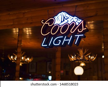 HAPPY VALLEY-GOOSE BAY, NEWFOUNDLAND AND LABRADOR, CANADA - JULY 26, 2019: Coors Light Beer Neon Sign With Rustic Canadian Wooden Cabin In The Background. Antlers Candelabra Hang On The Ceiling.