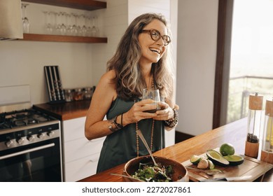 Happy vegan woman smiling while holding a glass of green juice in her kitchen. Mature woman serving herself organic food at home. Woman taking care of her aging body with a healthy plant-based diet.