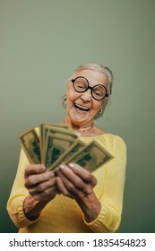 Happy Very Emotional Old Woman Counting Dollars, Holding Money And Laughing. Financial Literacy Concept. Round Glasses. Casual Clothes. Isolated Over Light Studio Background