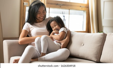 Happy Vietnamese millennial mom or nanny and little girl child relax on sofa in living room play together at home, smiling Asian young mother engaged in funny game enjoy weekend with small daughter