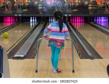 Happy View Of Young Girl Bowling With A Bowling Ramp Helping Her To Guide The Bowling Ball. A Family Activity Of Colorful, Cosmic Bowling Alley. Watching For A Strike.