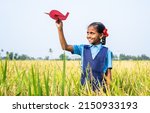 Happy village girl kid with school uniform playing using toy cardboard aeroplane at paddy field - concept of childhood dream, aspirations and freedom.