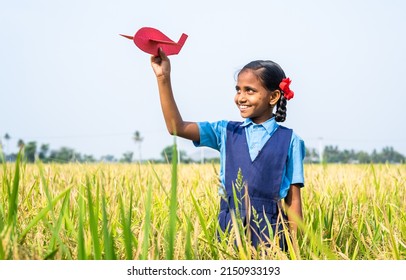 Happy village girl kid with school uniform playing using toy cardboard aeroplane at paddy field - concept of childhood dream, aspirations and freedom.