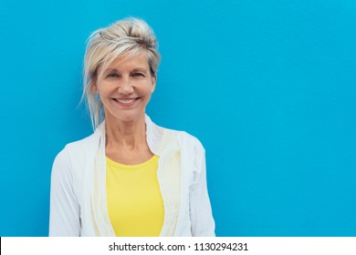 Happy vivacious older blond woman in a colorful yellow top posing against a bright blue wall with copy space