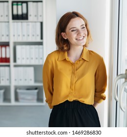 Happy vivacious young woman smiling to herself as she stands looking through a glass door at the office with a beaming smile