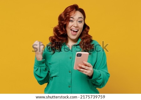 Happy vivid excited young ginger chubby overweight woman 20s wears green shirt hold in hand use mobile cell phone celebrate clenching fists say yes isolated on plain yellow background studio portrait