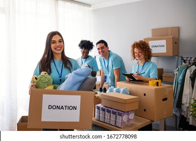 Happy Volunteer Looking At Camera. Woman Organizes Food And Clothing Drive. She Is Smiling At The Camera And Is Holding A Donation Box. Small Group Of People Working On Humanitarian Aid Project