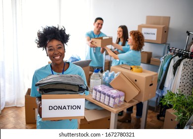 Happy Volunteer Looking At Camera. Woman Organizes Food And Clothing Drive. She Is Smiling At The Camera And Is Holding A Donation Box. Small Group Of People Working On Humanitarian Aid Project