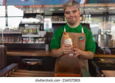 Happy Waiter With Down Syndrome Standing By Counter And Giving Sandwich To A Costumer In Cafe At Gas Station.