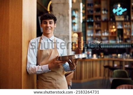 Happy waiter holding a tray while working in cafe and looking at camera.