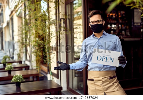 Happy waiter with protective face mask\
holding open sign while standing at cafe doorway.\
