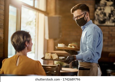 Happy Waiter Serving Food To A Guest While Wearing Protective Face Mask And Gloves In A Pub. 