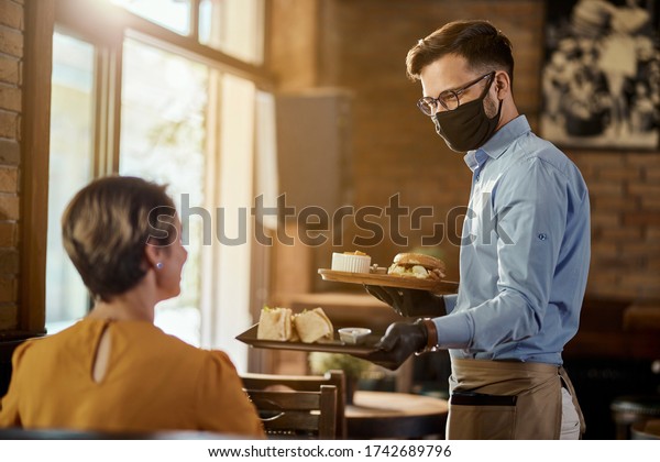 Happy waiter serving food to a\
guest while wearing protective face mask and gloves in a pub.\

