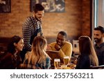 Happy waiter showing the menu on touchpad to group of guests in a bar.