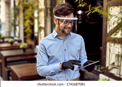 Happy waiter using digital tablet while wearing visor and protective gloves at outdoor cafe.  - Powered by Shutterstock