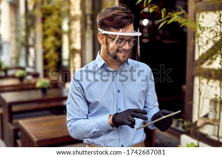 Happy waiter using digital tablet while wearing visor and protective gloves at outdoor cafe. 