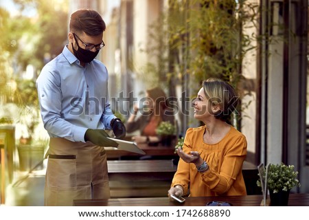 Happy waiter wearing protective face mask while showing menu on digital tablet to female guest in a cafe. 