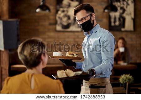 Happy waiter wearing protective face mask and gloves while bringing food to a customer in a pub. 