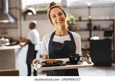 Happy waitress in a coffee shop holds a tray of delicious food, ready to serve customers and provide an excellent dining experience. Female server displaying a welcoming smile and a positive demeanour - Powered by Shutterstock