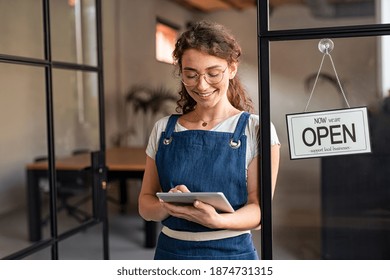 Happy waitress standing at restaurant entrance holding digital tablet to check the reservations. Smiling owner in blue apron standing at coffee shop entrance leaning on door with open signboard. - Powered by Shutterstock