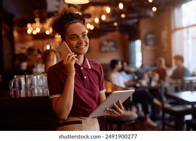 Happy waitress using touchpad while communicating on mobile phone in a bar.