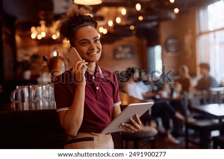 Happy waitress using touchpad while communicating on mobile phone in a bar.