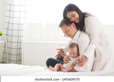 Happy And Warm Moment Of Asian Family Father And Mother Touching And Cuddling Their Cute Newborn Baby On Bed With White Curtains Background At Home. Complete Family Lifestyle Love And Care Concept