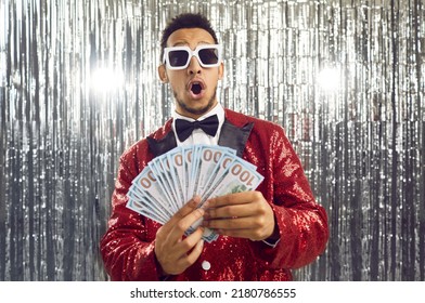 Happy wealthy ethnic winner man in shiny suit, bowtie and cool glasses looks at paper money bills bunch with surprised wow face expression. TV game show host guy in foil fringe studio shows prize cash - Shutterstock ID 2180786555