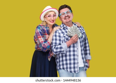 Happy Wealthy Family, Adult Man And Woman In Casual Checkered Shirt Standing Pickaback Together, Holding Fan Of Dollar, Toothy Smile, Looking At Camera. Indoor,isolated, Studio Shot, Yellow Background