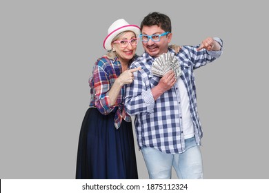 Happy Wealthy Family, Adult Man And Woman In Casual Checkered Shirt Standing Pickaback Together, Holding Fan Of Dollar And Pointing Finger To Money. Indoor,isolated, Studio Shot, Gray Background