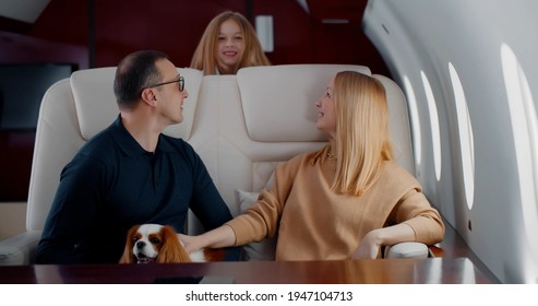 Happy Wealthy Family With Preteen Daughter And Cocker Spaniel Dog Flying Together On Private Jet. Mature Elegant Man And Woman Relaxing In Leather Chair With Little Daughter Jumping In First Class