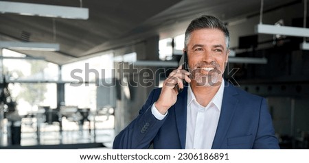Happy wealthy successful mature business man talking on the phone in office. Smiling professional businessman executive entrepreneur wearing suit holding telephone making corporate call on cellphone.