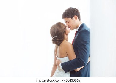 Happy Wedding Couple In Love. Beautiful Bride In White Dress With Brides Bouquet And Handsome Groom In Blue Suit Standing And Embracing Each Other And Kissing Indoors At Home Against Window, White