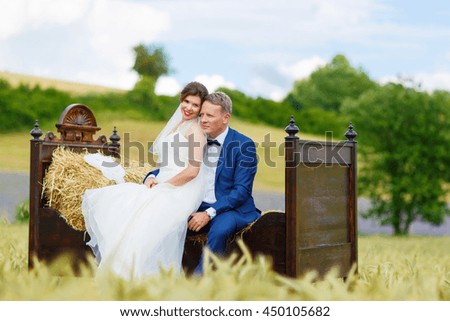Happy wedding couple in wheat field. Beautiful bride in white dress and groom having fun on bed in hay on summer day. Just married, young family.