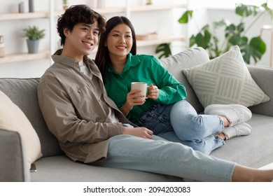 Happy Weekend Together. Affectionate Young Asian Couple Drinking Coffee And Cuddling On Cozy Couch At Home, Copy Space. Loving Millennial Spouses Enjoying Hot Beverage In Living Room