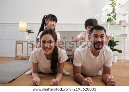 Happy well-being Asian Thai family, children play and tease their parents while yoga fitness training and health exercise together in the white living room, domestic home lifestyle, weekend activity.