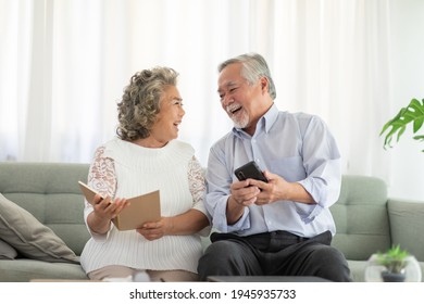 Happy Wellness Asian Elderly Or Seniors Couple Holding Mobile Phone And Book Smile And Laugh Together On Sofa In Living Room At Home,Retirement Lifestyle At Home Concept