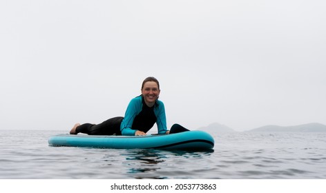 Happy wet woman sup surfer in wetsuit posing on board at water surrounded by fog