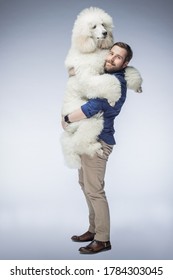 Happy White Guy Holds A White Poodle Puppy In His Arms. A Man Smiles At His Dog In The Isolated Background. The Dog And Its Owner.