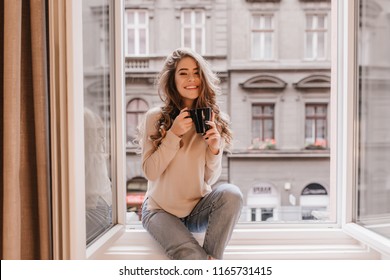 Happy white woman with gorgeous hairstyle sitting on window sill. Indoor portrait of glad young lady in jeans posing emotionally while drinking coffee. - Powered by Shutterstock