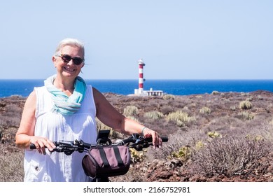 Happy White-haired Active Senior Woman Using Electric Bicycle At Sea, Elderly Caucasian Female In Summer Vacation, Lighthouse At The Horizon