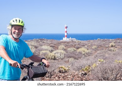 Happy White-haired Active Seniorman Cycling Off Road With Electric Bicycle At Sea, Elderly Caucasian Male In Summer Vacation, Lighthouse At The Horizon