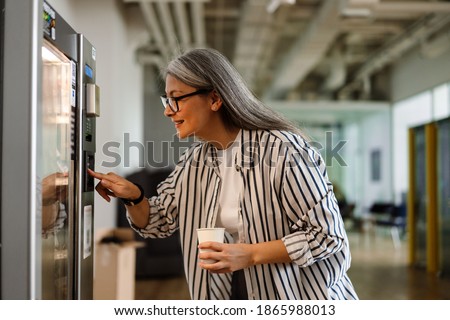 Happy white-haired mature woman using vending machine while drinking coffee indoors