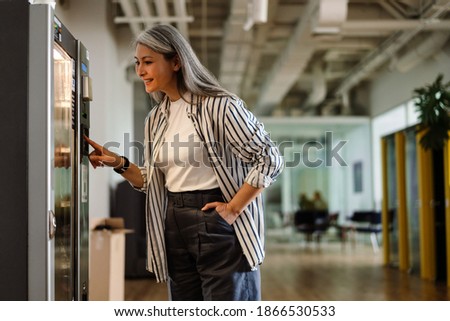 Happy white-haired mature woman using vending machine and smiling indoors
